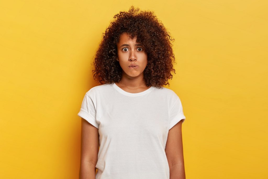 Worried lovely girl with bushy curly hair bites lips, has surprised nervous expression, feels embarrassed to speak in public place wears white wear poses over yellow background. Human emotions concept