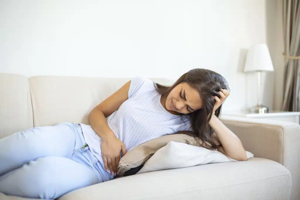 Woman in painful expression holding hands against belly suffering menstrual period pain,sitting sad on home bed, having tummy cramp in female health concept