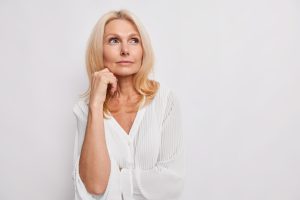 thoughtful blonde middle aged woman ponders on something keeps hand near face has healthy skin minimal makeup makes choice wears white blouse poses indoor blank copy space for your promotion