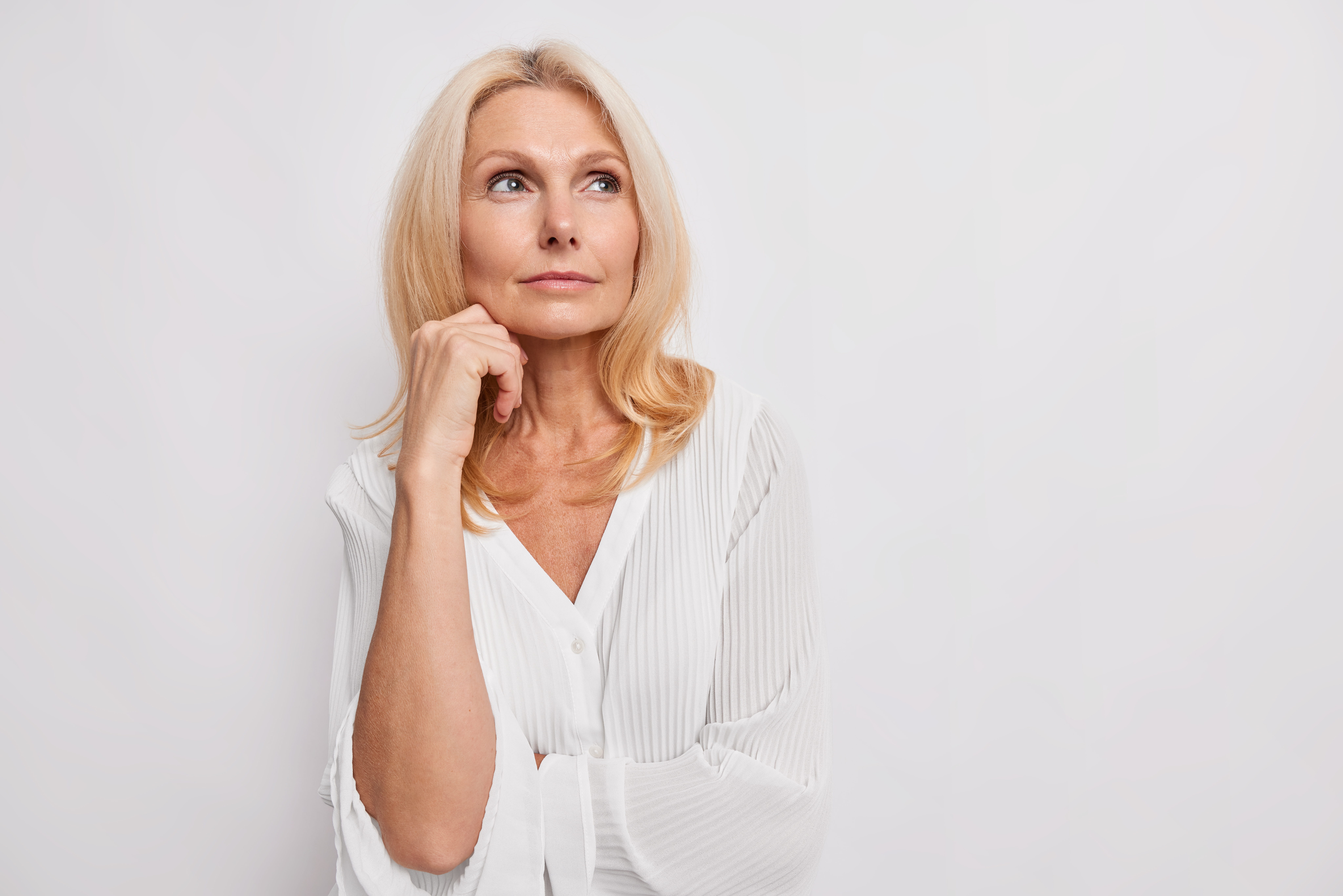 thoughtful blonde middle aged woman ponders on something keeps hand near face has healthy skin minimal makeup makes choice wears white blouse poses indoor blank copy space for your promotion