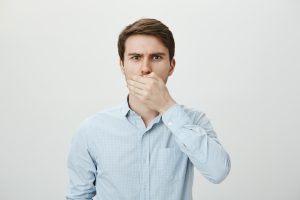 guy is gonna throw up. worried shocked caucasian male model covering mouth with palm, looking nervous while trying to keep secret, standing against gray background. man promised not to tell anything